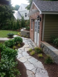 Stone pathway alongside brick building with white double doors, lanterns, plants, and lawn.