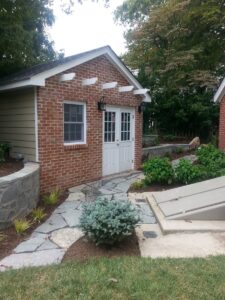 Small brick building with white double doors, window, stone path, plants, and concrete structure.