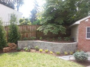 Stone retaining wall with shrubs and plants in a backyard beside a brick house and wooden fence.