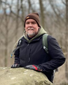 Man in brown beanie and black jacket outdoors with backpack, resting hands on large rock.