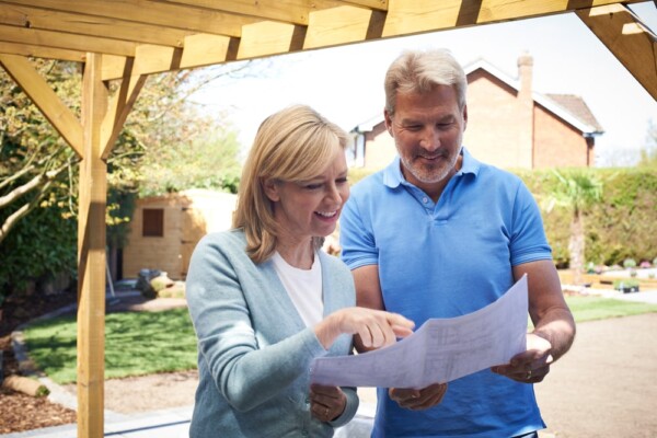 Man and woman reviewing plans under wooden pergola in backyard with lawn and brick house.