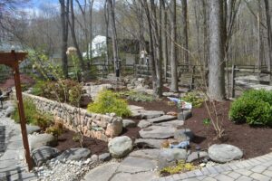 Stone pathway in landscaped garden with shrubs, a birdhouse, and wooden fence in early spring.