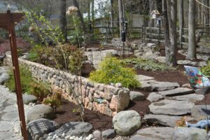 Landscaped garden with stone retaining wall, flagstone steps, shrubs, birdhouses, and wooden fence.