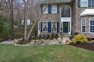 Stone house with blue shutters and door, front yard garden, rocks, and grassy lawn.