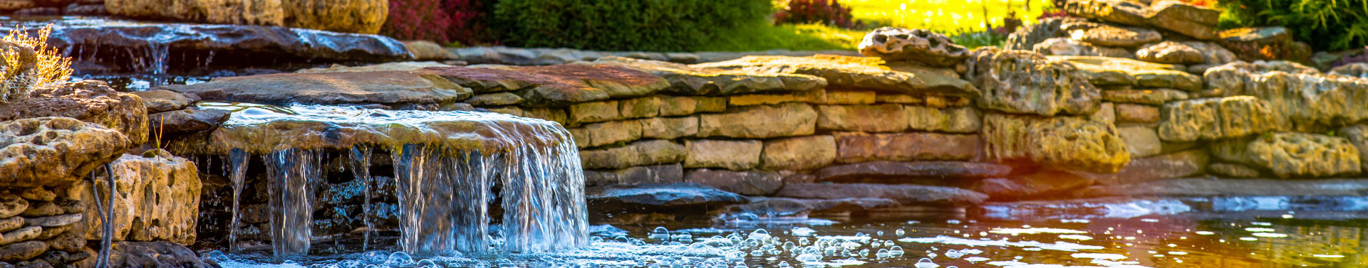 Stone waterfall with water flowing over rocks into a pond surrounded by greenery and foliage.