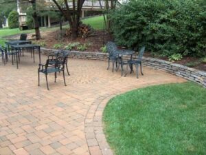 Paved patio with metal chairs and tables next to curved lawn and stone retaining wall with plants.