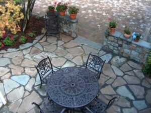 Outdoor stone patio with round metal table, four chairs, potted plants, and autumn tree.