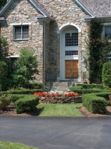 Stone house entrance with wooden door, arched window, stone stairs, and garden with flowers.