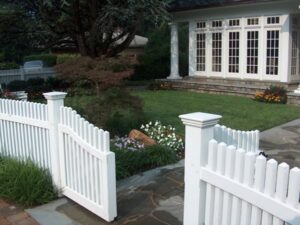 White picket fence with open gate leading to stone path, garden, and house porch with columns.