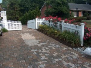 Interlocking paver driveway leading to a white picket fence with gate and flower garden.