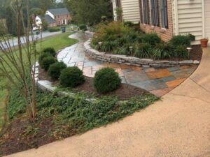 Residential front yard with multicolored stone walkway, curved retaining wall, and shrubs next to house.