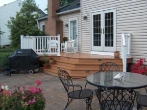 Backyard patio with glass-top metal table, four chairs, wooden steps, covered grill, and plants.