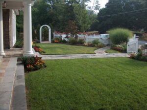 Front yard with green lawn, stone walkway, flower beds, white picket fence, and an arched gate.