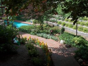 Brick pathway winds through garden with flowers, shrubs, tree, and a pool beside a stone wall.