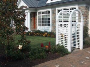 White wooden garden arbor with lattice design near brick pathway and house exterior with large windows and stone walls.