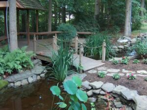 Wooden footbridge over a pond with plants, rocks, and a gazebo in a wooded garden setting