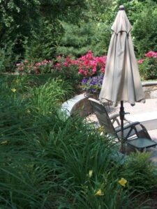 Patio chair with closed beige umbrella near stone border and colorful garden flowers.