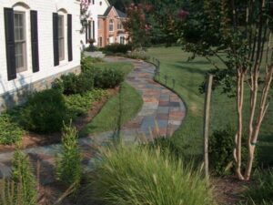 Stone pathway curves through garden beside white house with black shutters and lawn.