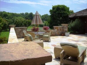 Outdoor stone patio with wicker chairs, a table, umbrella, and potted plants overlooking trees.