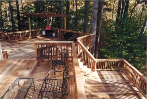 Multi-level wooden deck with metal chairs, tables, a BBQ grill under pergola, and surrounding trees.