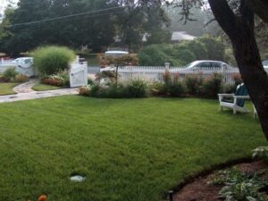 Front yard with green lawn, white picket fence with open gate, bushes, and a white chair.
