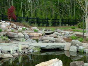 Landscaped garden with stone bridges over a small pond, surrounded by rocks, plants, and black fence.