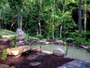 Small natural pond with rocks, plants, stone bench, and stepping stones in a green wooded area