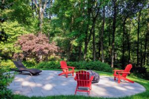 Backyard patio with stone paving, three red Adirondack chairs around covered fire pit, black lounge chair, surrounded by trees.