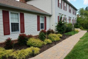 Two-story white house with red shutters, landscaped garden, paved walkway, and lawn.
