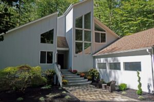 Modern two-story light gray house with large windows, curved steps, and a garage with patio seating.