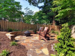 Backyard patio with stone floor, two wooden Adirondack chairs, covered fire pit, curved stone wall, wooden fence, and surrounding greenery.