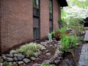 Narrow water stream with rocks and plants beside brick building and wooden bridge over water.