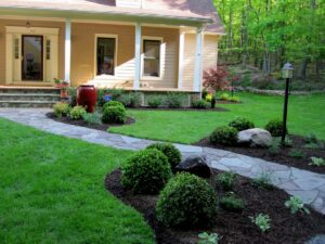 Beige house with porch, stone pathway, lawn, decorated garden beds, and lamp post.