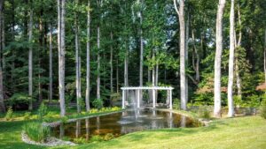 Landscaped garden with pond, central water fountain, green grass, tall trees, and white pergola seating.
