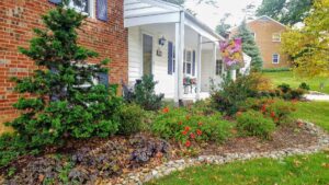 Front of a brick and white house with blue shutters, porch, garden, and decorative flag.