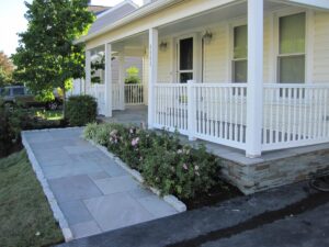 Front porch of light-colored house with white railings, stone siding, paved walkway, and pink flowering shrubs.