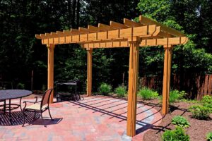 Wooden pergola on a paved patio with metal table, chair, grill, and surrounding garden beds.