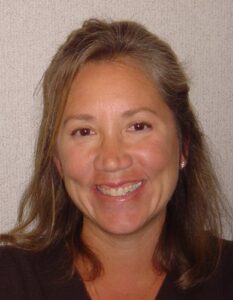 Smiling woman with shoulder-length light brown hair wearing a dark top against a textured background