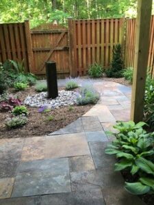 Backyard with stone tile pathway, circular garden bed with rocks, plants, and tall black sculpture.