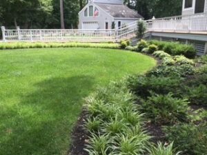 Curved garden edge with trimmed grass, plants, white fence, and light-colored house.