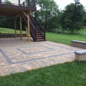 Backyard with tiled patio, low stone wall, raised wooden deck, stairs, grass, and trees.