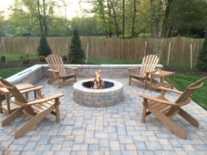 Outdoor stone fire pit with five wooden Adirondack chairs on a paved patio surrounded by trees and a wooden fence