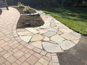 Backyard with brick and stone paved pathway, a garden bed with plants, and surrounding grass.