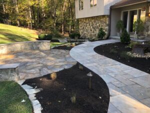 Stone pathway and patio with garden beds and landscaped bushes outside a house entrance.