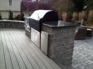 Outdoor kitchen with stone island, covered grill, stainless steel cabinets, wooden deck, and stone patio.