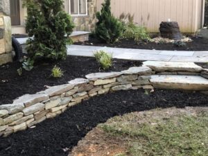 Curved stone retaining wall with steps, dark mulch, plants, paved walkway, and grass area.