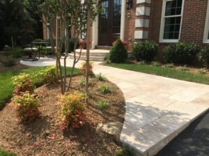 Paved walkway to brick house entrance with landscaped garden, small plants, tree, and patio seating.