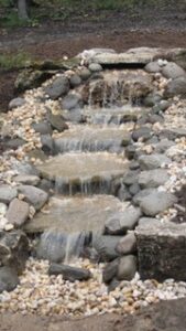 Small man-made waterfall flowing over stone steps, bordered by rocks and gravel.