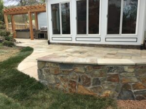 Backyard patio with stone retaining wall, tiled floor, wooden pergola, and outdoor grill.