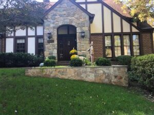 Stone and brick house entrance with dark door, stone wall, bushes, flowers, and trees.
